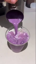 Hand pouring vibrant purple ube mixture from measuring cup into glass with ice to make an ube latte
